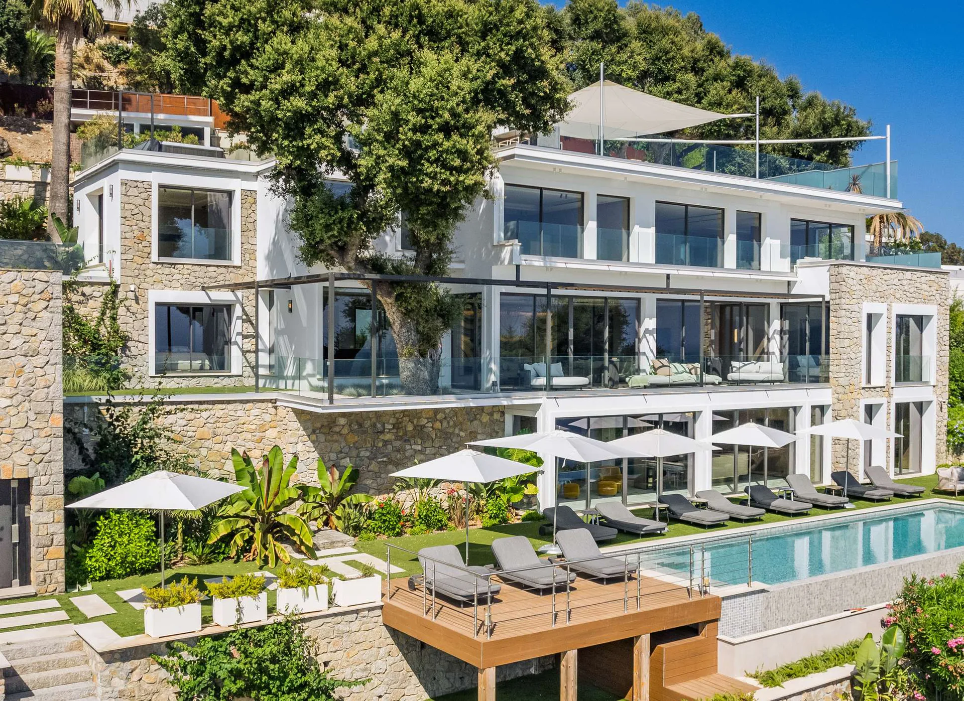 Piscine vue de côté avec cyprès, parasols et façade pierre